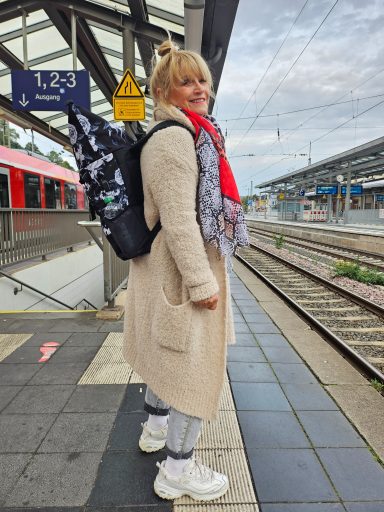 Frau mit Rucksack und Schal steht am Bahnsteig, wartet auf den Zug.
