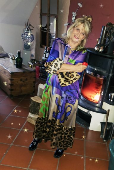 Frau in einem bunten, gemusterten Kleid steht vor einem Kamin in einem gemütlichen Raum.