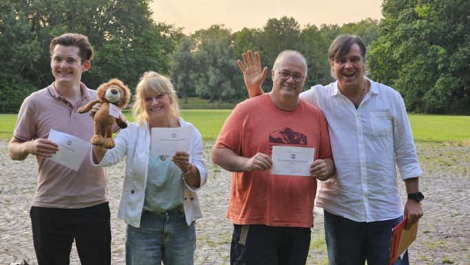 Vier Personen stehen lächelnd im Freien, eine mit einem Stofftier, alle halten Karten.