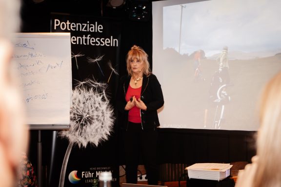 Frau spricht vor einer Präsentation, mit Flipchart und Projektor im Hintergrund.