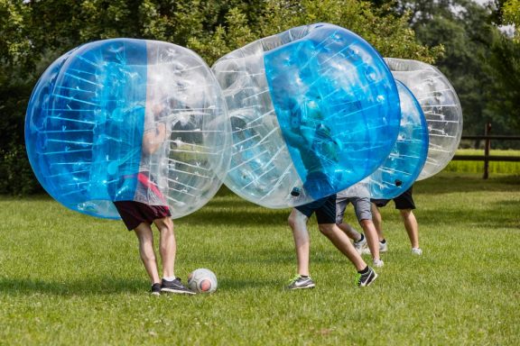 Drei Personen in aufblasbaren Bällen spielen auf einer Wiese Fußball.