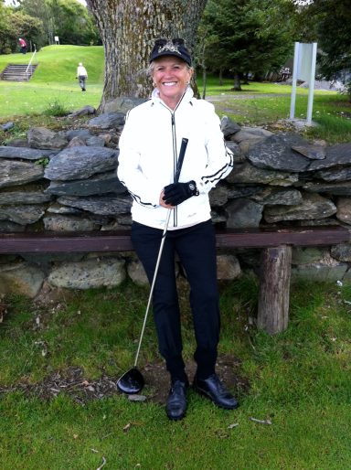 Eine Person mit Golfschläger steht vor einer Steinmauer in einer grünen Umgebung.