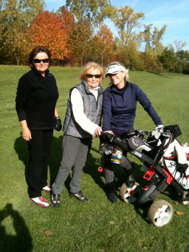 Drei Frauen auf einem Golfplatz, eine mit Golfwagen, umgeben von herbstlichen Bäumen.