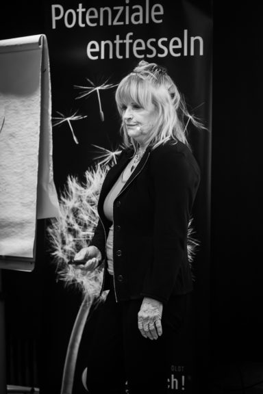 Frau mit langen, blonden Haaren steht neben einem Flipchart vor einem Banner.